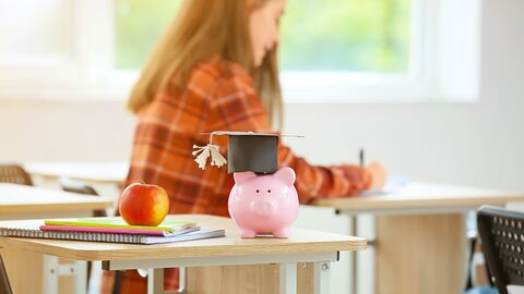 Bild zum Thema Ein rosa Sparschwein mit Absolventenhut auf einer Schülerbank im Unterricht