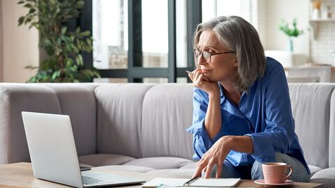 Bild zum Thema Eine Frau mittleren Alters sitzt zuhause auf dem Sofa und recherchiert an ihrem Notebook