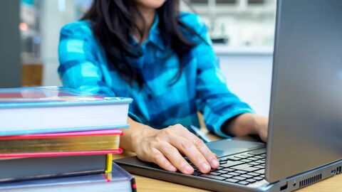 Eine Lehrerin zuhause vor ihrem Notebook; neben ihr ein Bücherstapel 