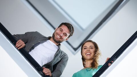 Bild zum Thema Ein angehender Lehrer und eine angehende Lehrerin, die im Treppenhaus einer Schule stehen und auf den Betrachter hinabschauen