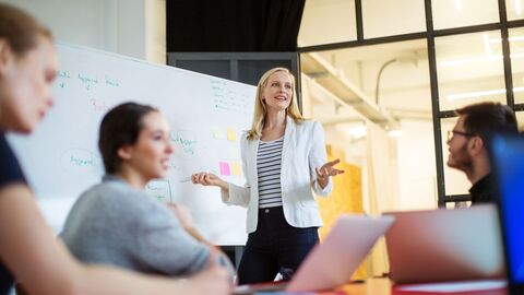 Bild zum Thema Eine Trainerin mit einer Lerngruppe im Gespräch; sie steht an einem Whiteboard