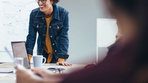 Eine Teamerin in flotter Jeansjacke während eines Workshops; sie stützt sich mit ihren Händen auf den Tisch auf und lächelt