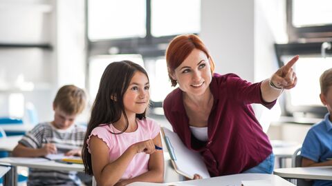 UBUS-Einstiegsfortbildung Eine Lehrerin erklärt einer Schülerin etwas an der Tafel; sie beugt sich zum Kind hinunter