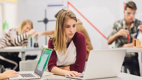 Fachtag Naturwissenschaften Eine Schülerin, die im Nawi-Unterricht konzentriert an einem Notebook arbeitet; im Hintergrund Mitschüler bei Experimenten