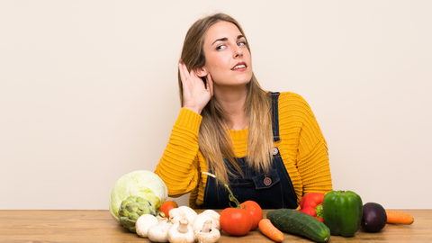 Schulverpflegung Eine junge Frau, die ihre Hand an ihr Ohr legt, um besser zu hören; vor ihr auf dem Tisch liegen Obst und Gemüse