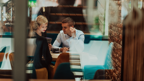 Eine Frau und ein Mann im Gespräch bei der Arbeit; man sieht sie durch eine Glasscheibe