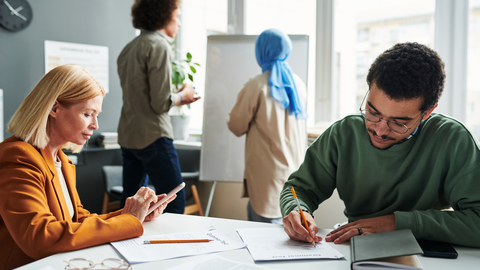 Fortbildung für UBUS-Kräfte Eine Lehrerin und eine UBUS-Kraft arbeiten zusammen; im Hintergrund arbeiten Schüler
